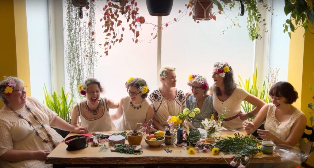 Women in white dresses and flower garlands sit around a table